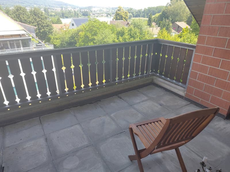 Balcony with a view of a lake and surrounding greenery, featuring a wooden chair and decorative railing.