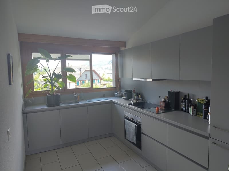 Modern kitchen with grey cabinets, tiled floor, and a large window with a potted plant on the windowsill. The kitchen features a sink, oven, coffee maker, and various bottles and containers on the countertops.