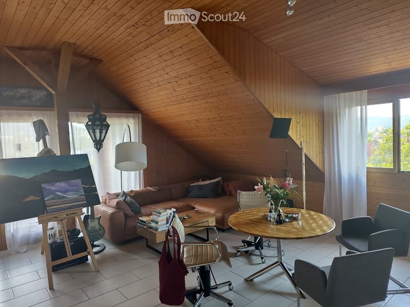 Cozy living room with wooden beams, a brown leather sofa, a round wooden coffee table, and a guitar on a stand. The room features large windows with white curtains and a view of the outdoors.