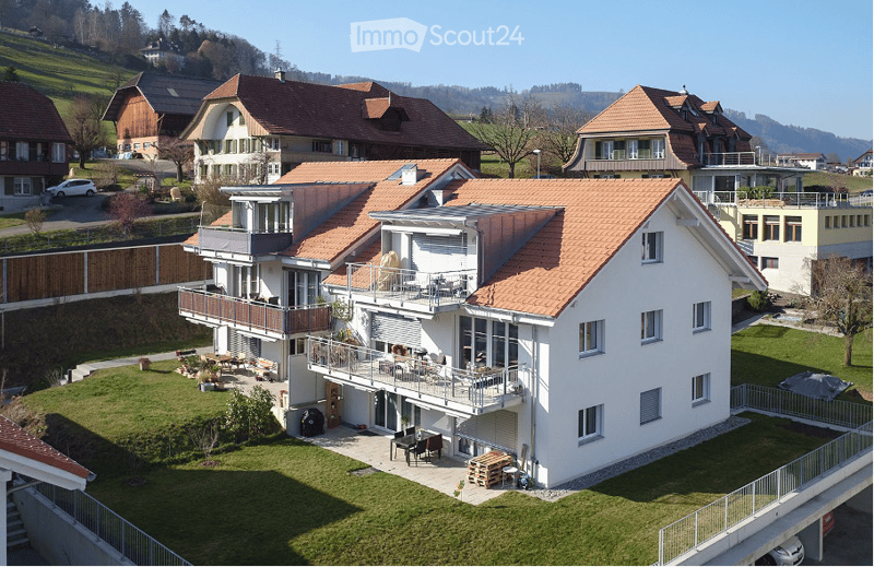 A modern two-story house with a red-tiled roof, white exterior, and multiple balconies. The house is surrounded by a well-maintained lawn and features outdoor seating areas and a patio.