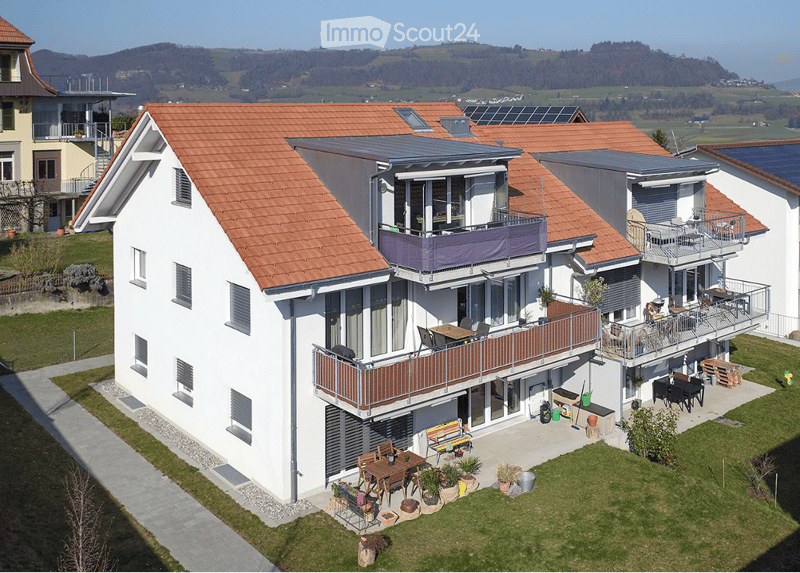 Modern two-story house with red-tiled roof, white exterior, multiple balconies, outdoor seating areas, and a scenic countryside view.