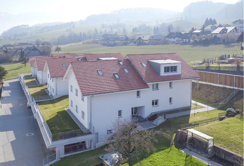 Aerial view of a modern two-story house with a red-tiled roof, white exterior, and a visible garage. The house is surrounded by a well-maintained lawn and is part of a residential neighborhood with other houses and open fields in the background.