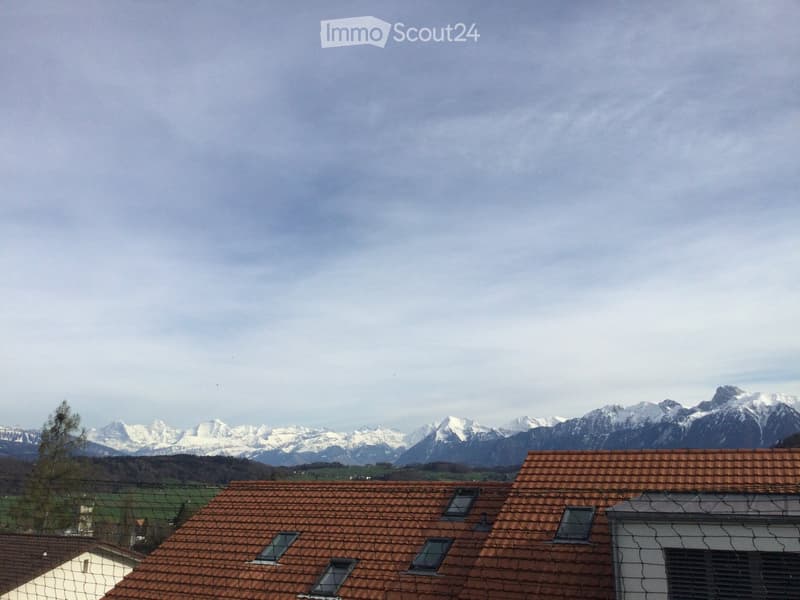 Roof tops of buildings with red-tiled roofs, surrounded by a scenic mountain range under a cloudy sky.