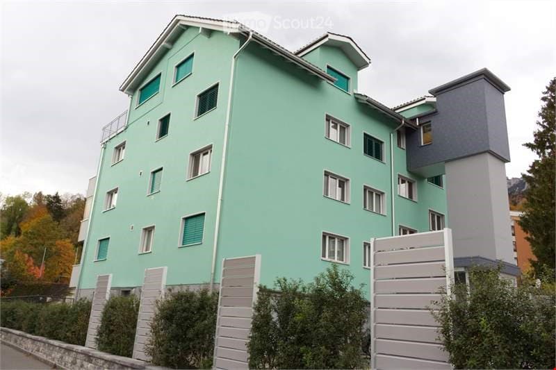 A modern residential building with three floors and several windows. The walls are painted green, and the roof has a triangular shape. The exterior is adorned with multiple white railings on the balcony, and there are bushes and trees on the sides. The bu