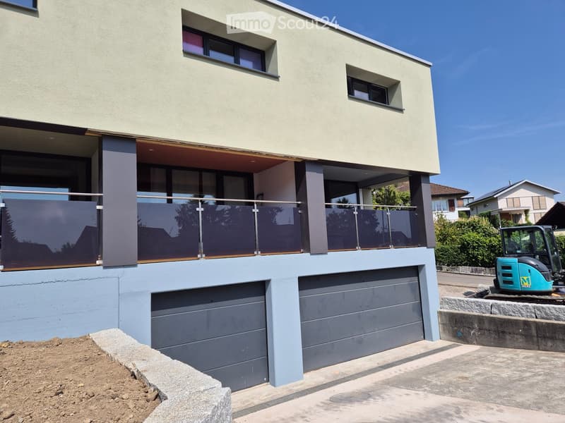 Modern two-story house with light-colored exterior, large windows, and a balcony on the upper level. Features a garage and raised ground floor.