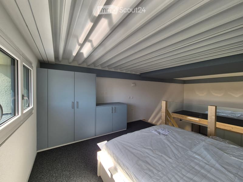 Modern, compact bedroom with a single bed, built-in wardrobe, desk, and chair. The room features a slanted ceiling and large windows allowing natural light.