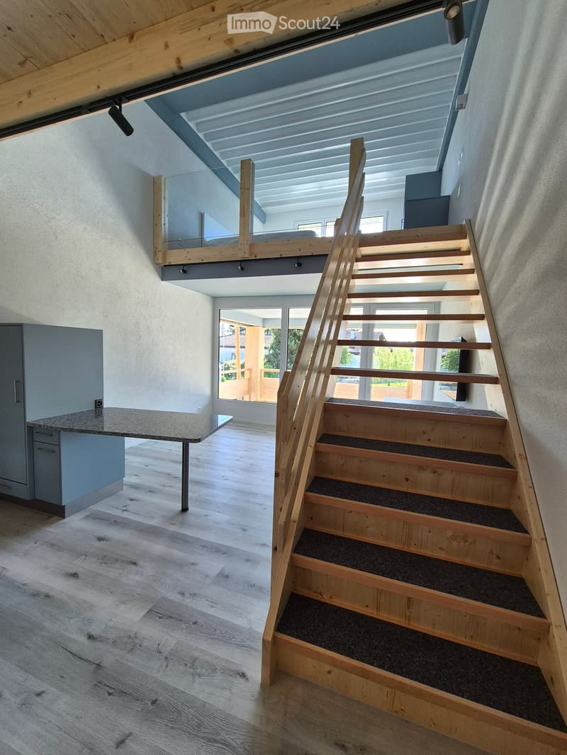 Modern hallway with wooden staircase, light wood flooring, and a small table with a countertop. The staircase has a wooden railing and carpeted steps, leading to an open loft area with a glass railing and skylight above.