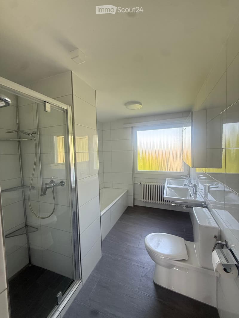 Modern bathroom with a bathtub, shower, toilet, double sink, and large window letting in natural light.