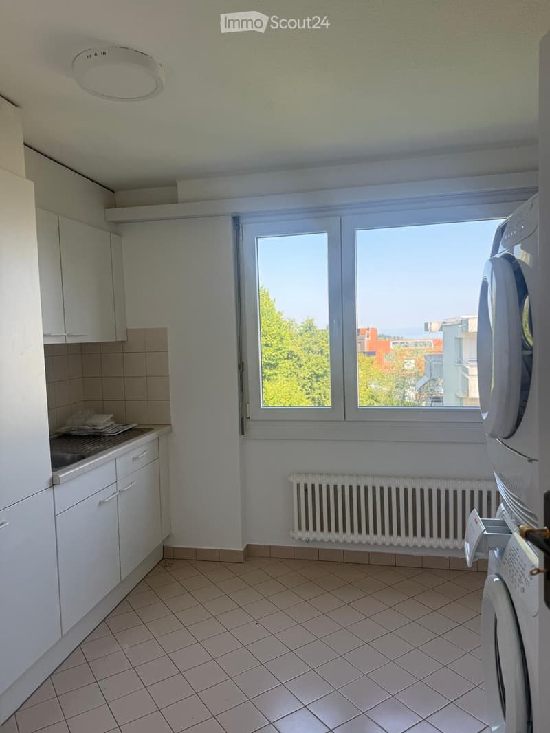 A small, well-lit room with tiled flooring, white cabinets, a sink, a radiator, and a washing machine. The room has large windows offering a view of greenery and buildings.
