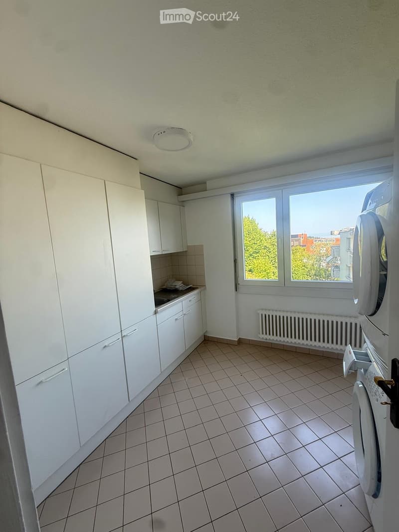 A small, unfurnished kitchen with white cabinets, tiled floor, and a window overlooking a cityscape. A washing machine is positioned near the window.