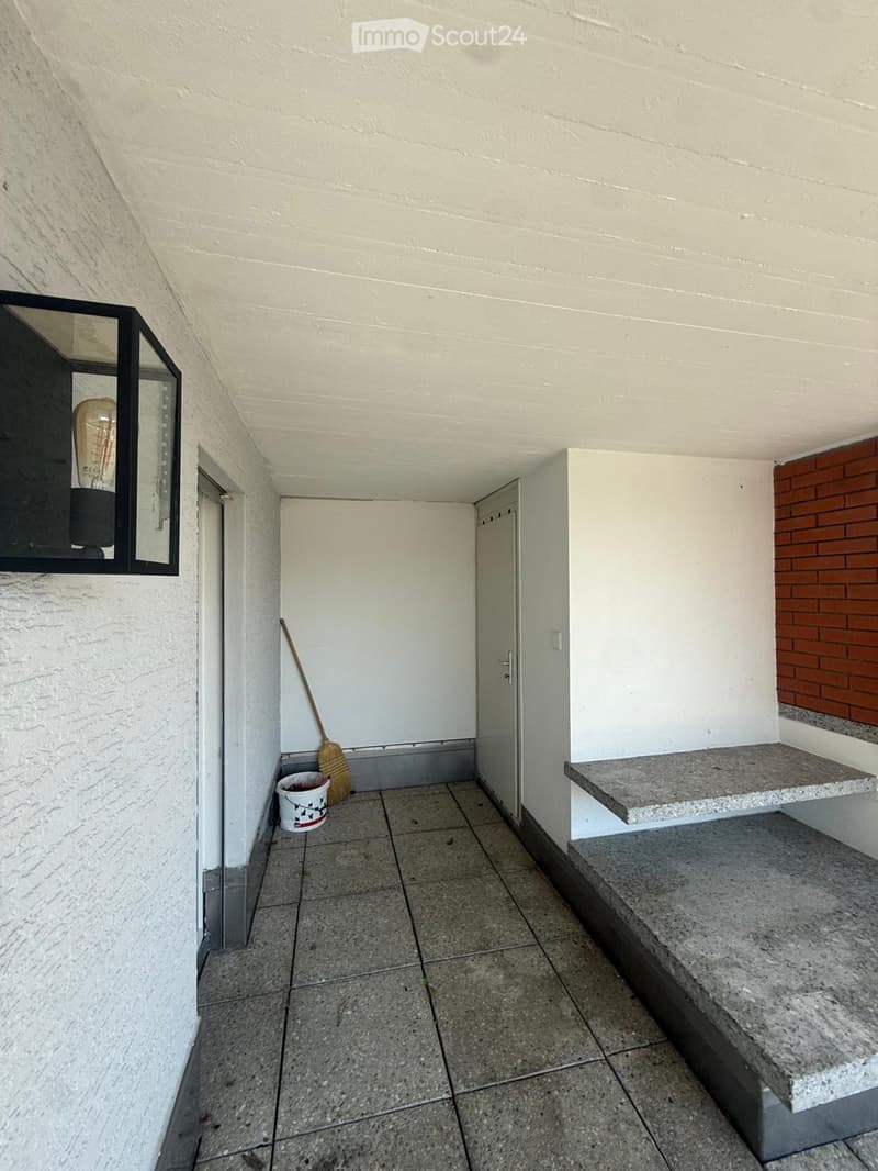 Empty hallway with tiled floor, white walls, a broom, and a small bucket near the entrance. A wall-mounted light fixture is visible on the left side.