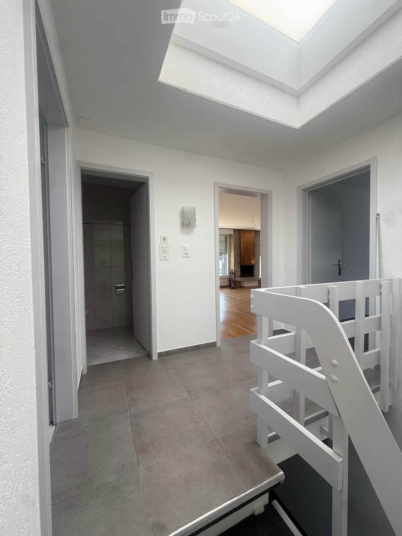 Bright hallway with tiled floor, white walls, and a skylight at the top. Features a white staircase railing on the right and three doors leading to different rooms.