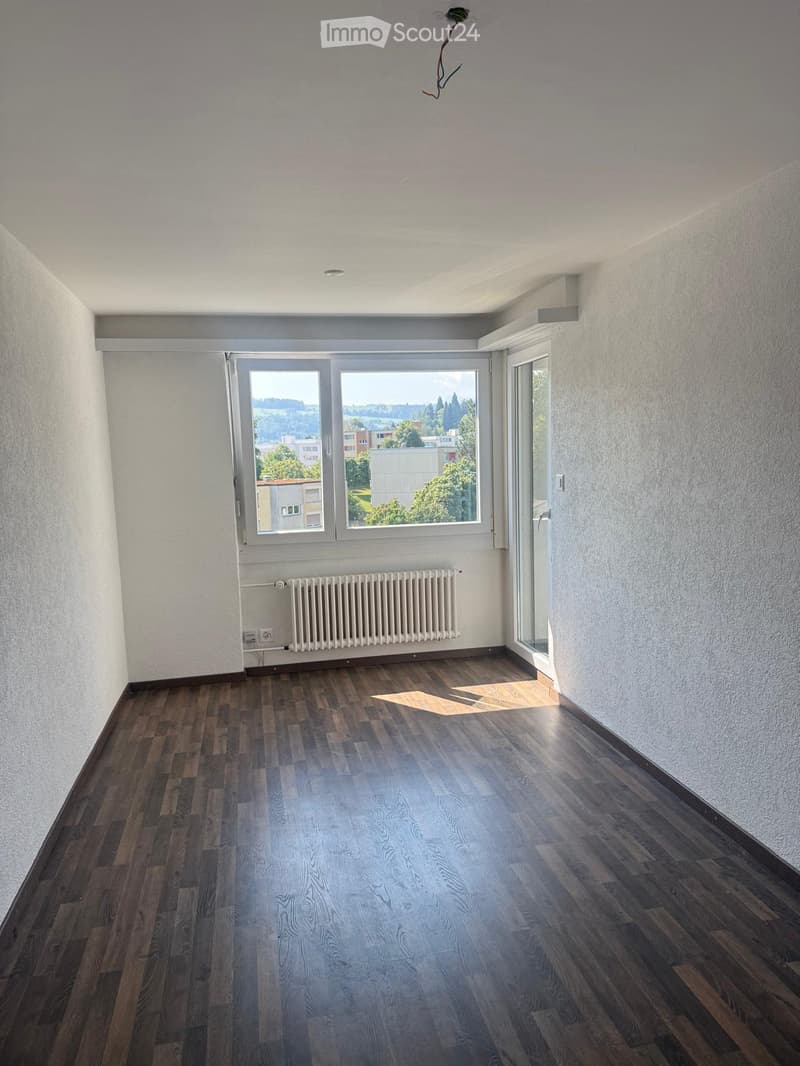 Empty room with wooden flooring, white walls, large window with view, radiator, and exposed electrical wiring on the ceiling.