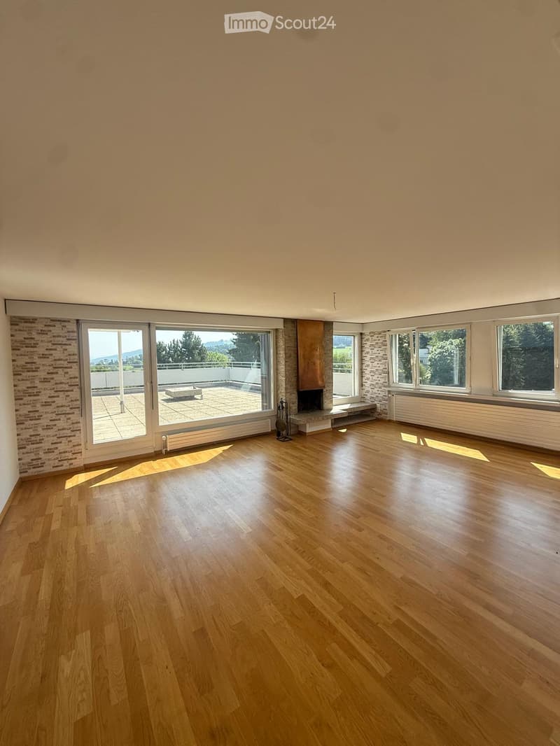 Empty room with wooden flooring, large windows, and a fireplace. The room has a balcony accessible through large glass doors.