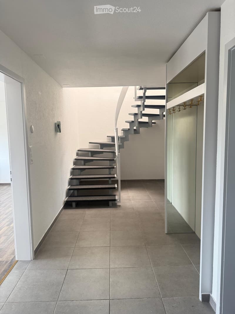 Modern hallway with tiled floor, white walls, and a staircase leading upwards. Features built-in shelving and coat hooks on the right side.