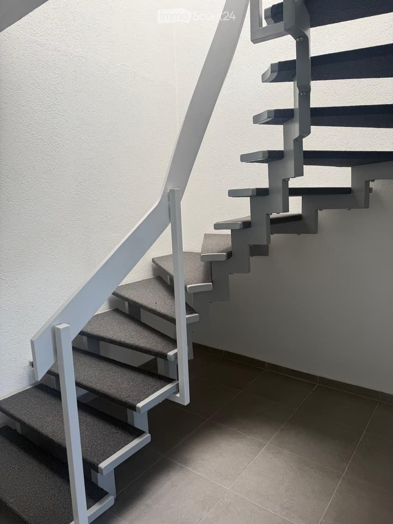 Modern staircase with gray carpeted steps and white metal railings, leading upwards against a plain wall.