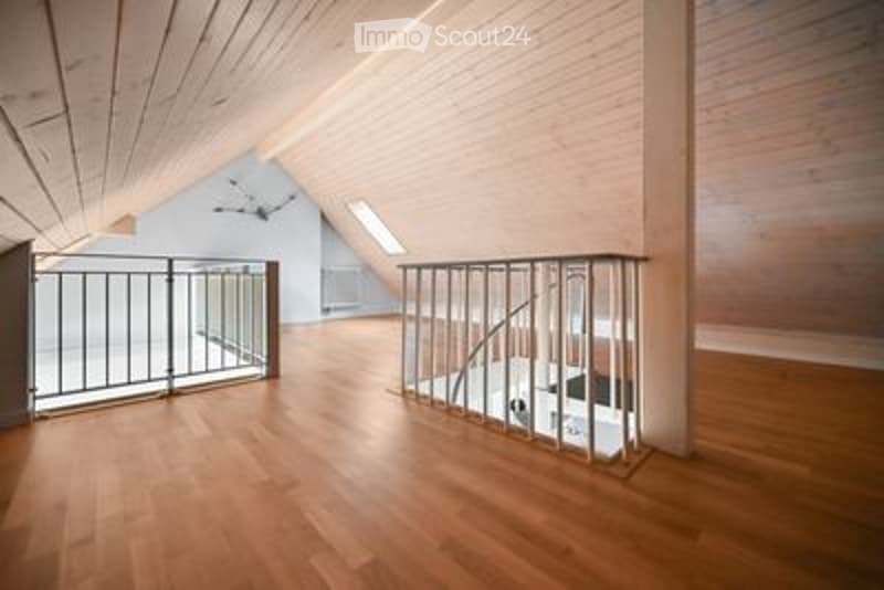 Empty room with wooden flooring and exposed wooden beams on the ceiling. A metal staircase leads to an upper level, and a small window is visible in the background.