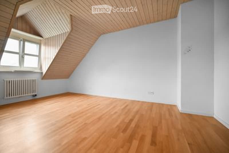 Completely empty room with wooden flooring, white walls, and a slanted wooden ceiling. A window with white frames and a radiator are visible on one wall.