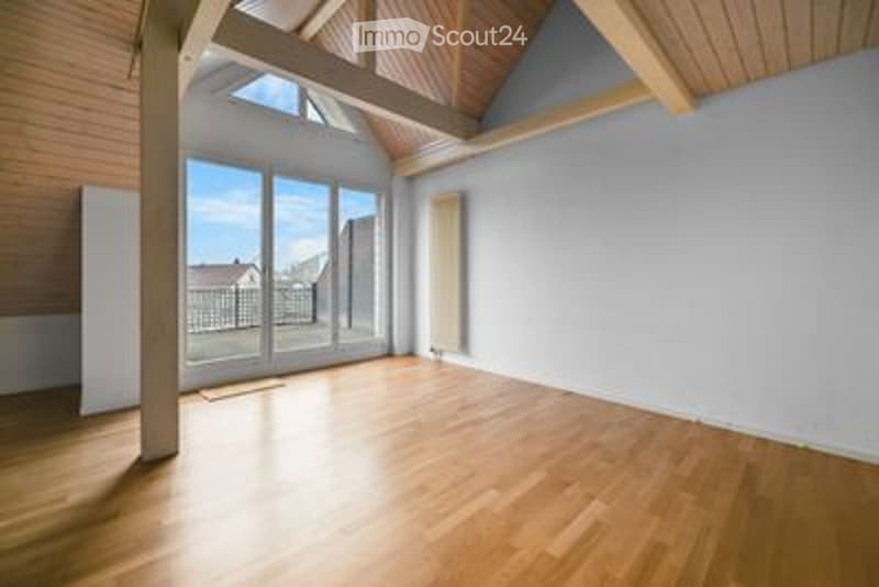 Bright, empty room with wooden flooring, large windows, and exposed wooden beams on the ceiling. The room has white walls and a view of the outdoors through the windows.