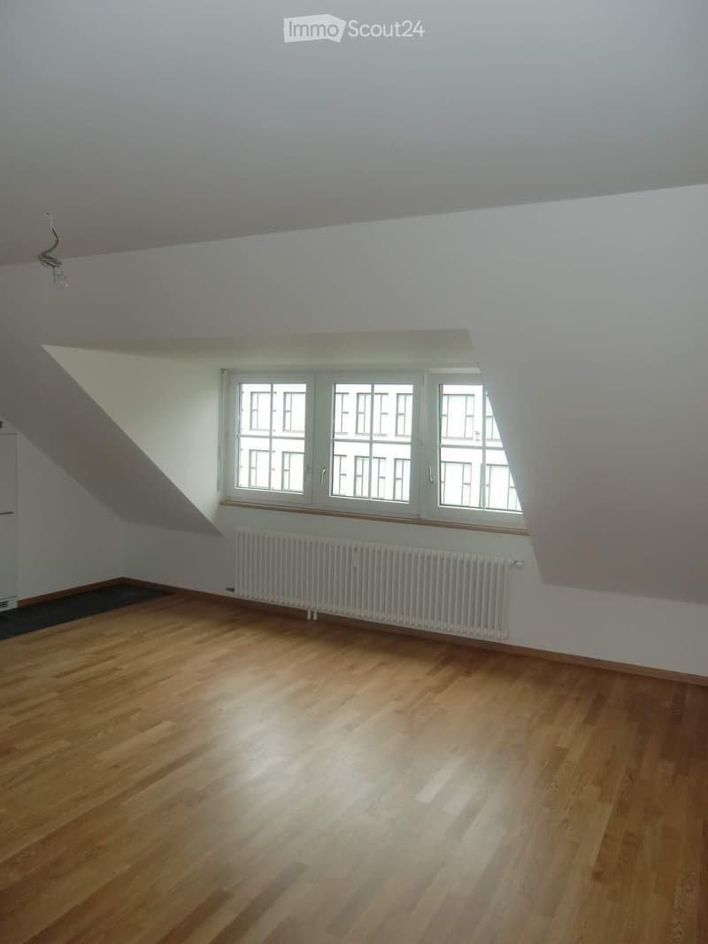 Empty room with wooden flooring, white walls, and large windows with white frames. A radiator is visible below the windows.