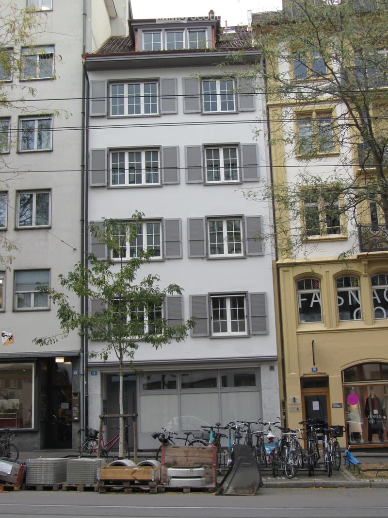 A three-story residential building with white facade and grey shutters, featuring multiple windows and a storefront at the ground level. Bicycles are parked in front, and there are some wooden crates and construction materials on the sidewalk.