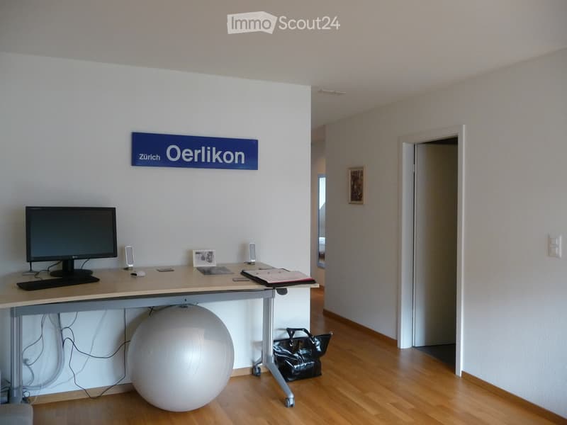 Office space with a desk, computer, keyboard, mouse, exercise ball, and handbag. A sign reading "Zürich Oerlikon" is on the wall.