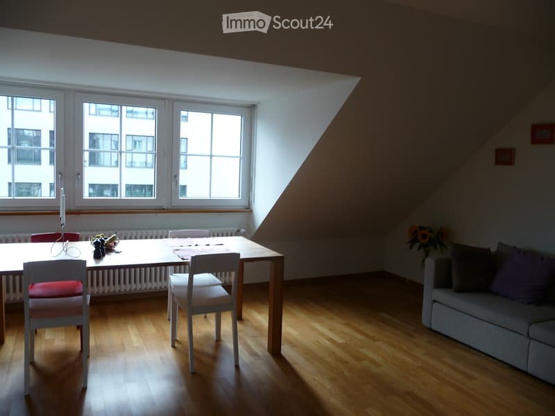 Bright, open living and dining area with wooden flooring, large windows, a grey sofa, a wooden dining table with chairs, and a vase of sunflowers.