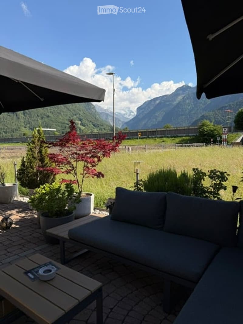 Outdoor seating area with a grey sofa, wooden coffee table, and several potted plants, overlooking a scenic mountain view.