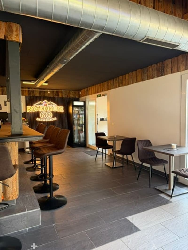 Modern restaurant interior with bar seating, tables, chairs, and a refrigerator. Exposed wooden walls and industrial-style lighting.