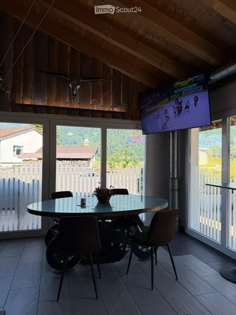 Bright room with large windows offering a view, featuring a round glass table surrounded by chairs, a television mounted on the wall, and a flower vase on the table.