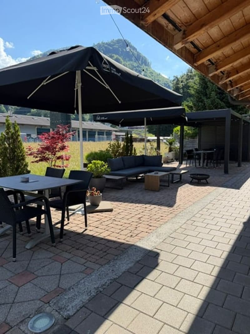 Outdoor patio area with a large umbrella, seating area including a sofa and coffee table, dining table with chairs, and potted plants. The area is paved with bricks and features a scenic mountain view in the background.