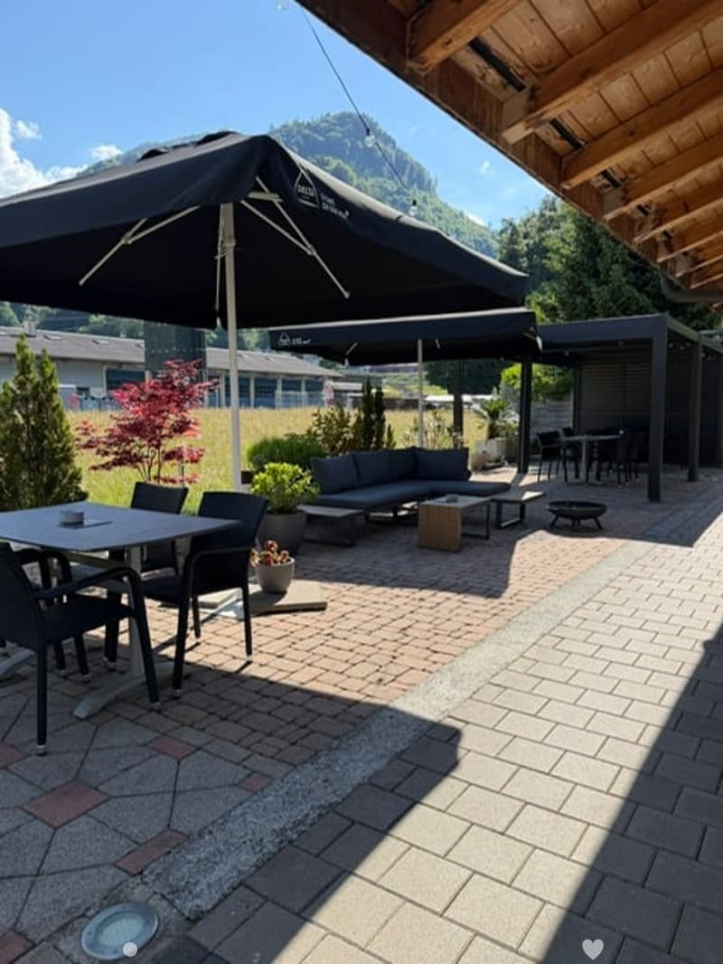 Outdoor patio area with a large umbrella, seating area including a sofa and coffee table, dining table with chairs, and potted plants. The area is paved with bricks and features a scenic mountain view in the background.