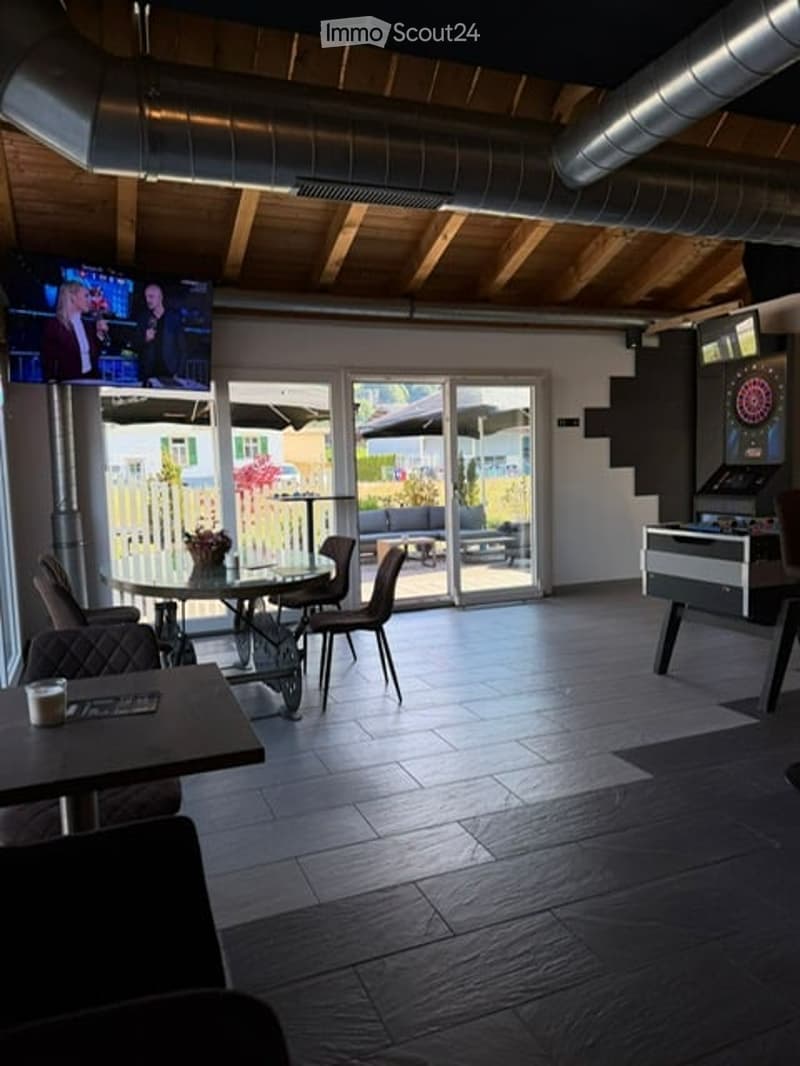 Modern lounge area with wooden ceiling beams, large glass doors leading to an outdoor patio, a round table with chairs, a sofa, a dartboard, and a screen displaying content.