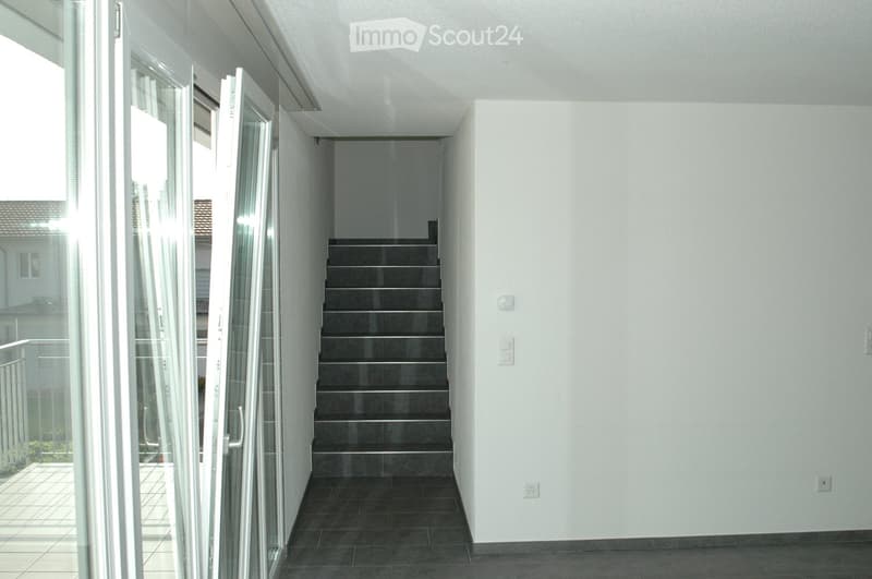 Modern hallway with a staircase leading upwards, white walls, dark tiled floor, and large glass sliding doors opening to a balcony area.