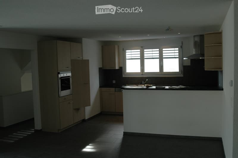 Modern kitchen with light wood cabinets, stainless steel oven, sink, and range hood. Features a central island with a black countertop and built-in storage. Natural light from three windows illuminates the space.