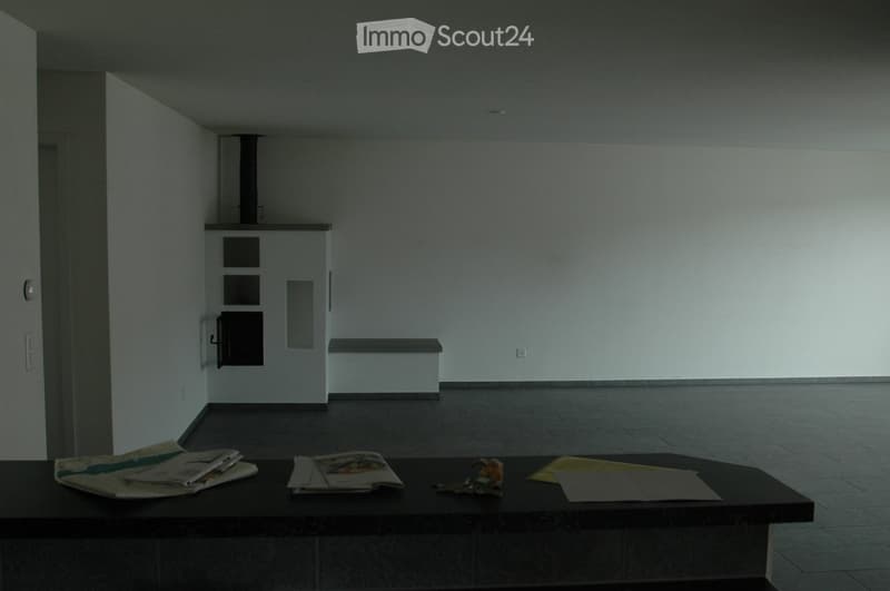 A small, unfurnished room with a dark wooden table holding magazines, a toy, and some papers. The room has white walls, a tiled floor, and a built-in bench along one wall.