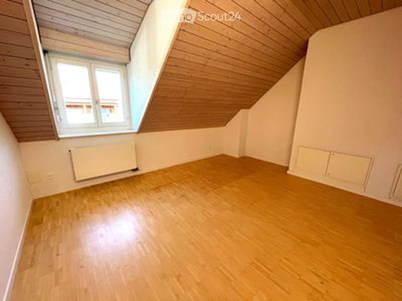 Completely empty room with wooden floor, slanted wooden ceiling, and a single window with white frame. No furniture or equipment.