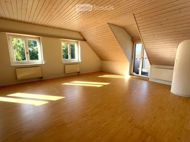 Bright, empty attic room with wooden floors and slanted wooden ceiling. Three windows provide natural light, and there are radiators along the walls.