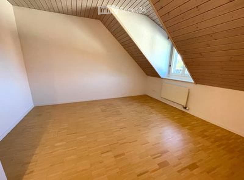 Empty room with wooden flooring, white walls, and a slanted wooden ceiling. A single window and a radiator are visible.