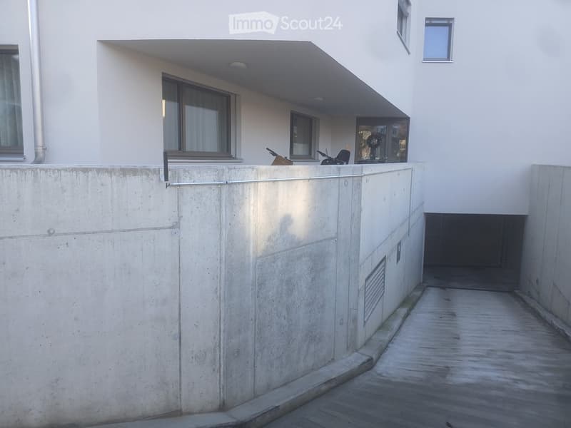 Modern building exterior with raised ground floor, concrete walls, and a narrow wooden walkway leading to an entrance.
