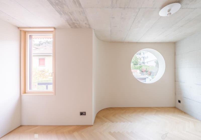 Empty room with light-colored walls, wooden floor, and two windows (one rectangular and one round).