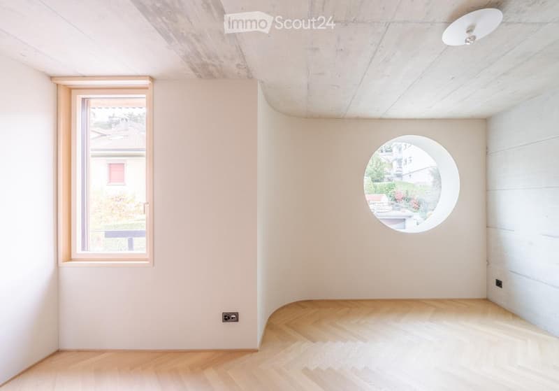 Empty room with light-colored walls, wooden floor, and two windows (one rectangular and one round).