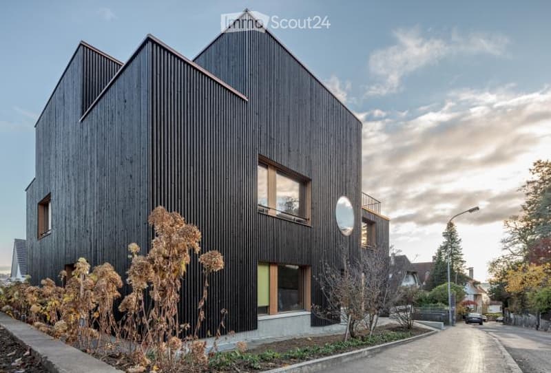 Modern two-story house with dark wooden facade, large windows, and a rounded window on the upper level. The house is surrounded by a small garden area with bare trees and shrubs.