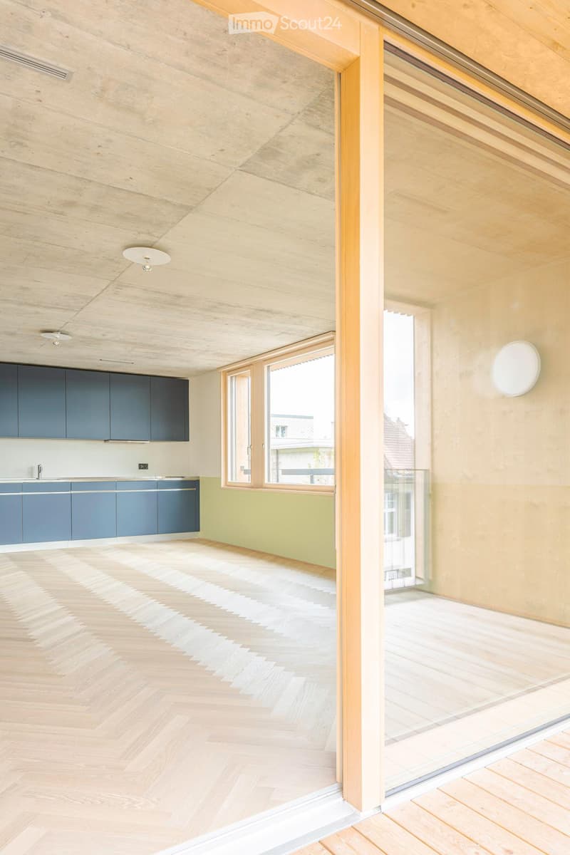 Open-plan space with light wooden flooring, grey cabinets, and large windows offering a view of the outside. The room is unfurnished, with only built-in cabinetry and a sink visible.