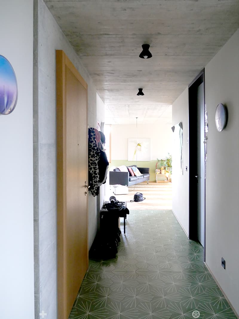 Modern hallway with geometric patterned tiles, recessed lighting, and a coat rack with jackets. The hallway leads to a living area with a sofa, colorful cushions, and a small table with plants.