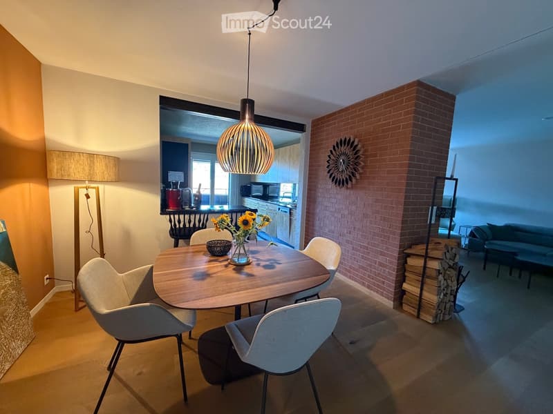 Modern dining area with a round wooden table, four chairs, a vase with sunflowers, and a bowl. Adjacent living area features a sofa, a table lamp, and a decorative wooden stack. Exposed brick wall and pendant light visible.