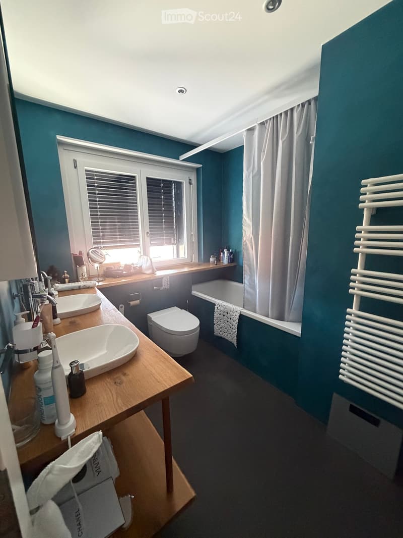 Modern bathroom with teal walls, double sink vanity, toilet, bathtub, radiator, and window with blinds.