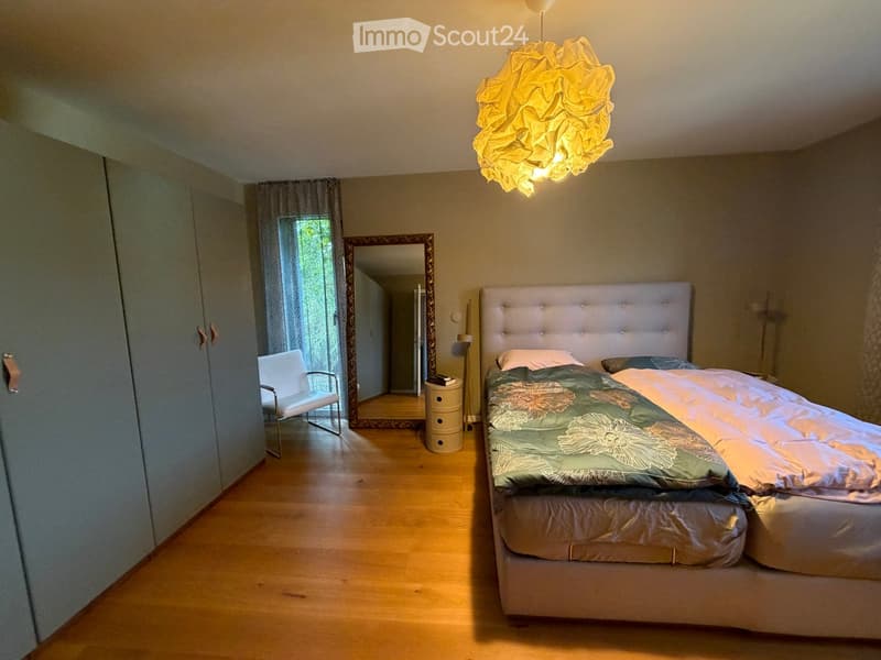 Well-lit bedroom with wooden flooring, a large upholstered headboard bed, a white chair, a mirror, and a decorative hanging lamp.