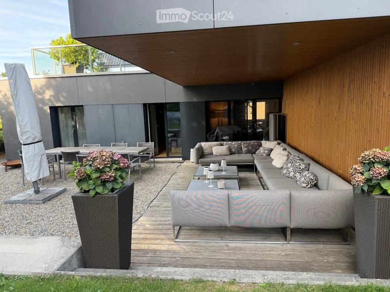 Modern outdoor patio with wooden decking, grey sectional sofa, coffee tables, dining area, and large umbrella.