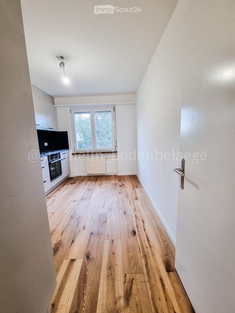 Empty hallway with wooden flooring, white walls, and a door on the right side. A small window with frosted glass is visible at the end of the hallway.